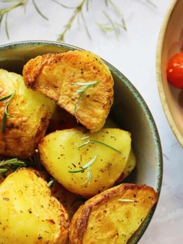 Roast potatoes topped with sprigs of rosemary in a bowl. A bowl of tomatoes sits on the side.